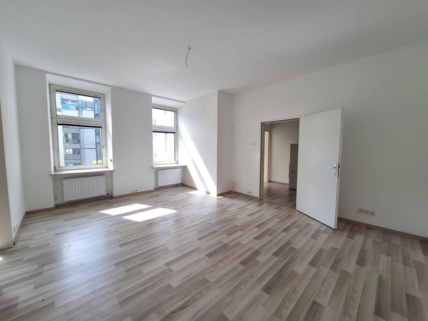 Geräumiges Wohnzimmer mit hellem Laminatboden und viel Tageslicht durch die Fenster.