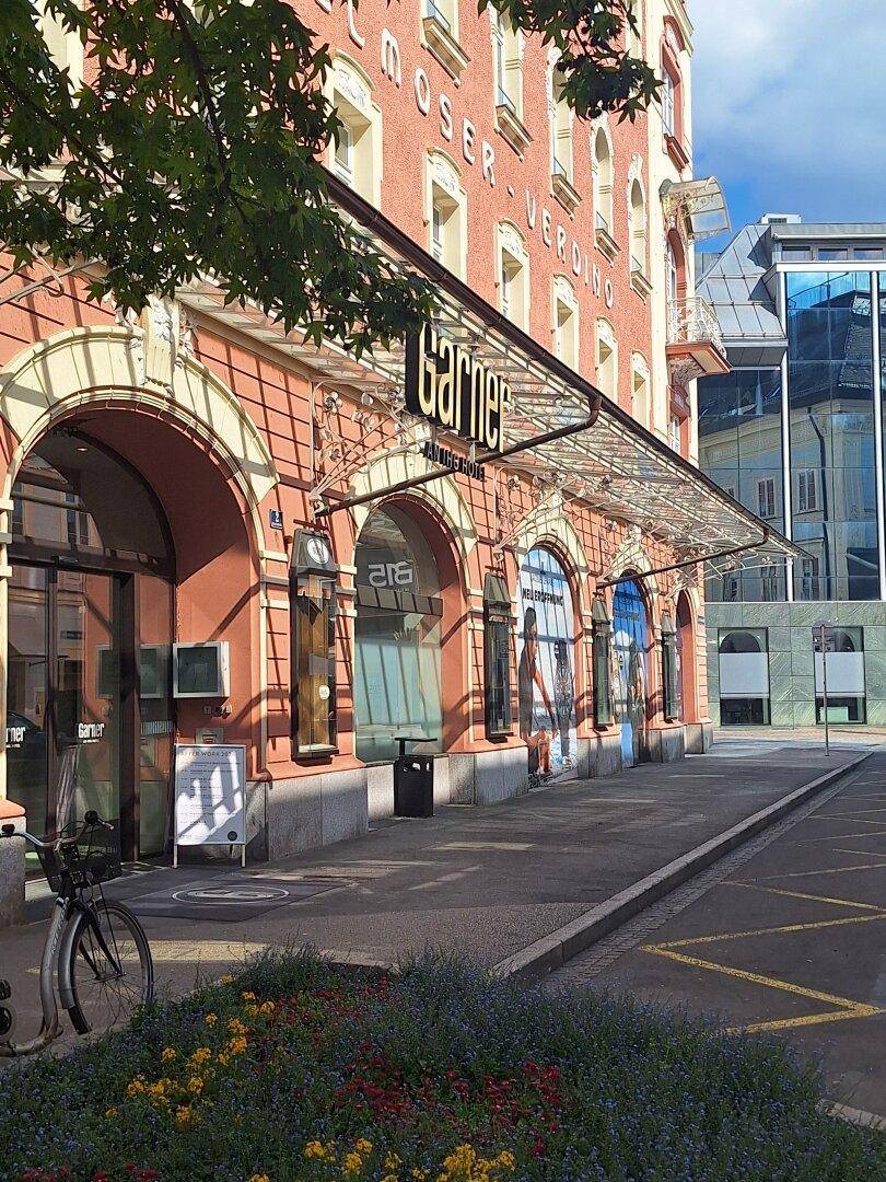 Historisches Gebäude mit Geschäften im Erdgeschoss, einem Glasvordach und einem Blumenbeet am Gehweg.