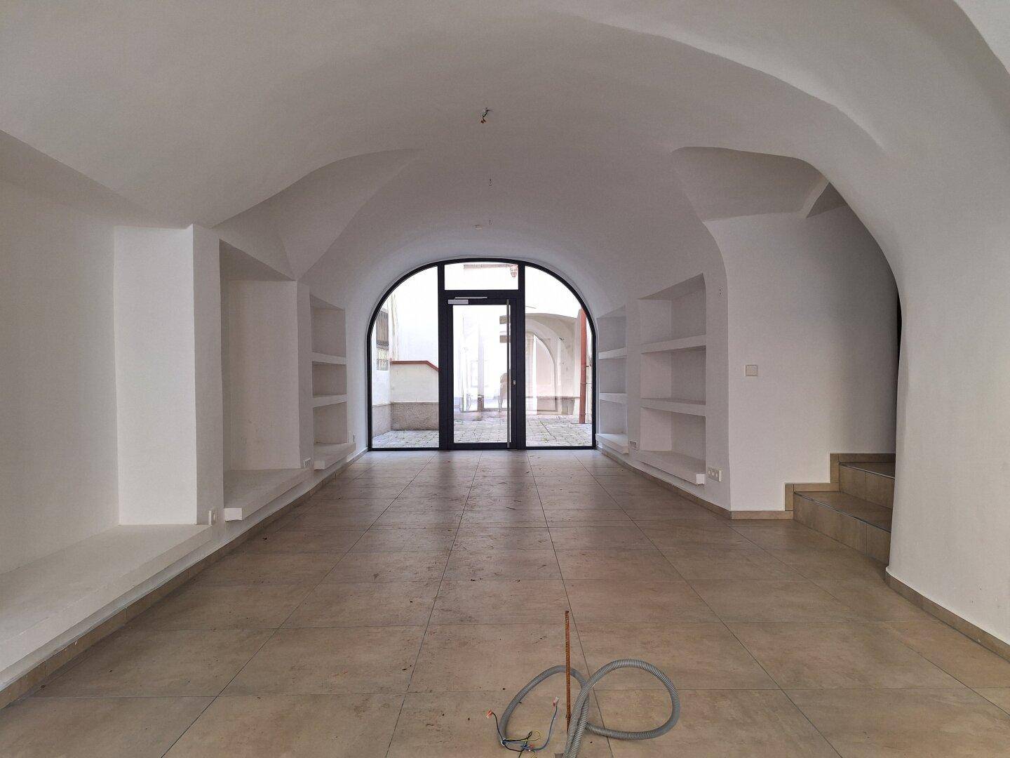 Geschäftsraum mit Gewölbedecken und Fliesenboden, der durch eine große Bogentür zum Innenhof führt.