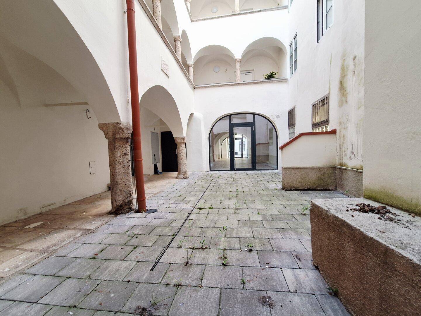 Blick über den gepflasterten Innenhof mit Arkaden und einem modernen Eingangsbereich.