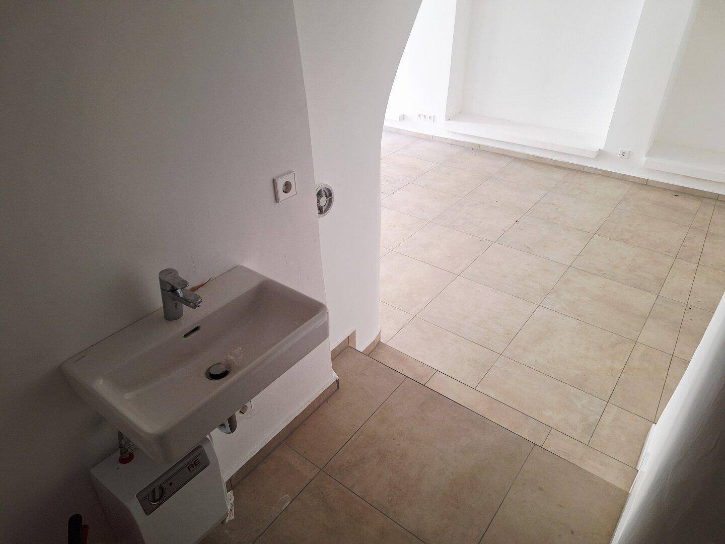 Einfacher Sanitärbereich mit Waschbecken und Fliesenboden, angrenzend an einen größeren Raum.