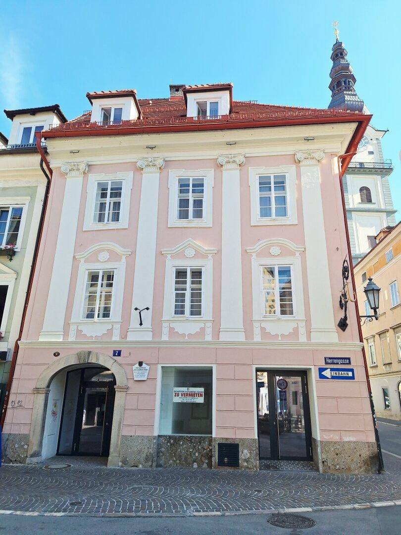 Historisches Gebäude mit rosa Fassade, weißen Fensterrahmen und einem Uhrturm im Hintergrund.