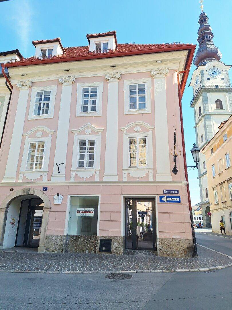 Historisches Gebäude mit rosa Fassade an einer Straßenecke, mit Uhrturm im Hintergrund.