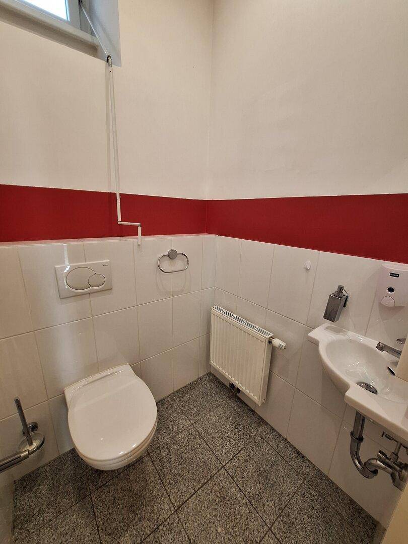 Kleines Gäste-WC mit weißen Fliesen, rotem Wandstreifen, Toilette und kleinem Waschbecken.