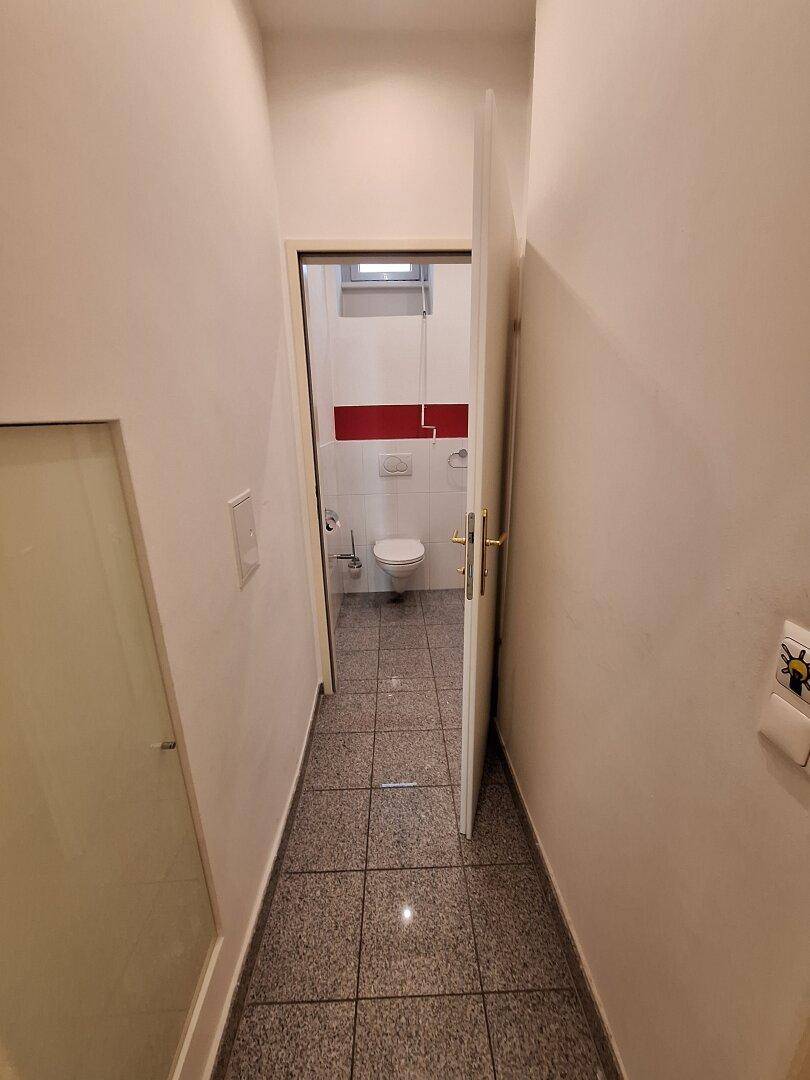 Schmaler Flur mit grauen Fliesen, der zu einem Badezimmer oder Gäste-WC führt.