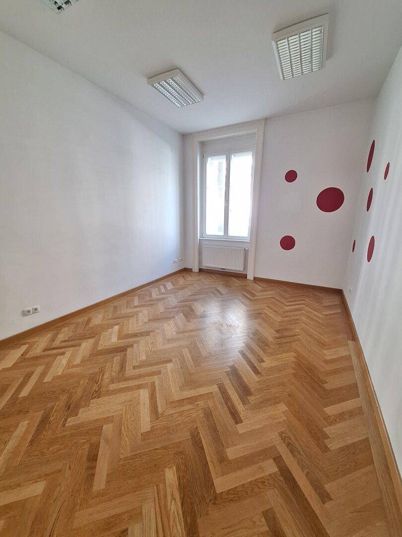 Leerer Raum mit Fischgrätparkett, weißen Wänden und roten Dekorpunkten sowie einem Fenster.