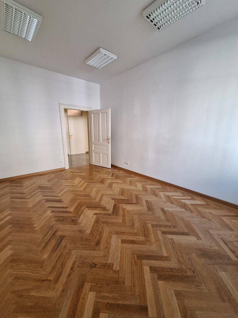 Geräumiger Raum mit Fischgrätparkettboden und einer offenen Tür, die den Blick in ein weiteres Zimmer freigibt.