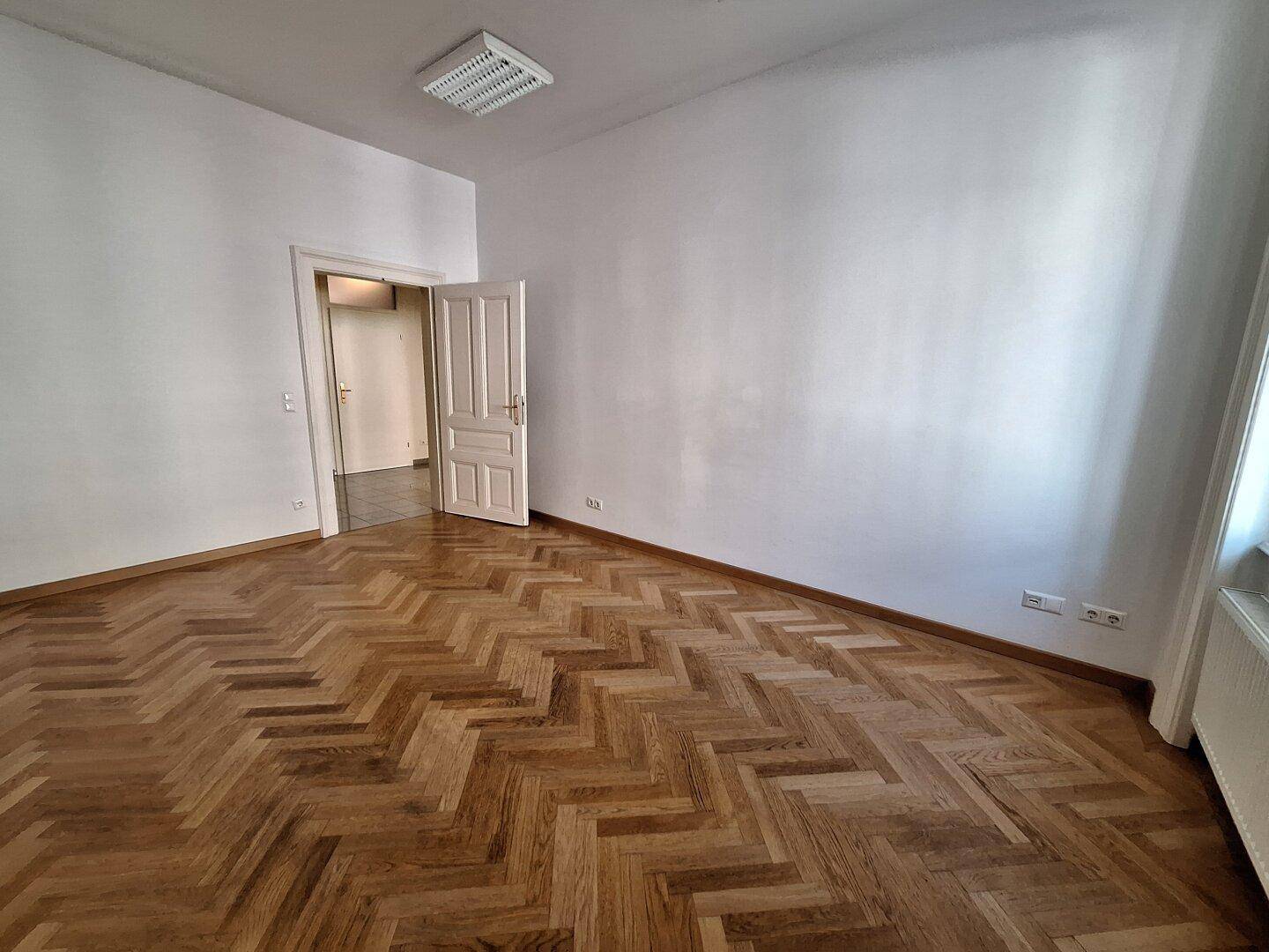 Heller Raum mit klassischem Fischgrätparkett und einer offenen Tür zu einem weiteren Zimmer.