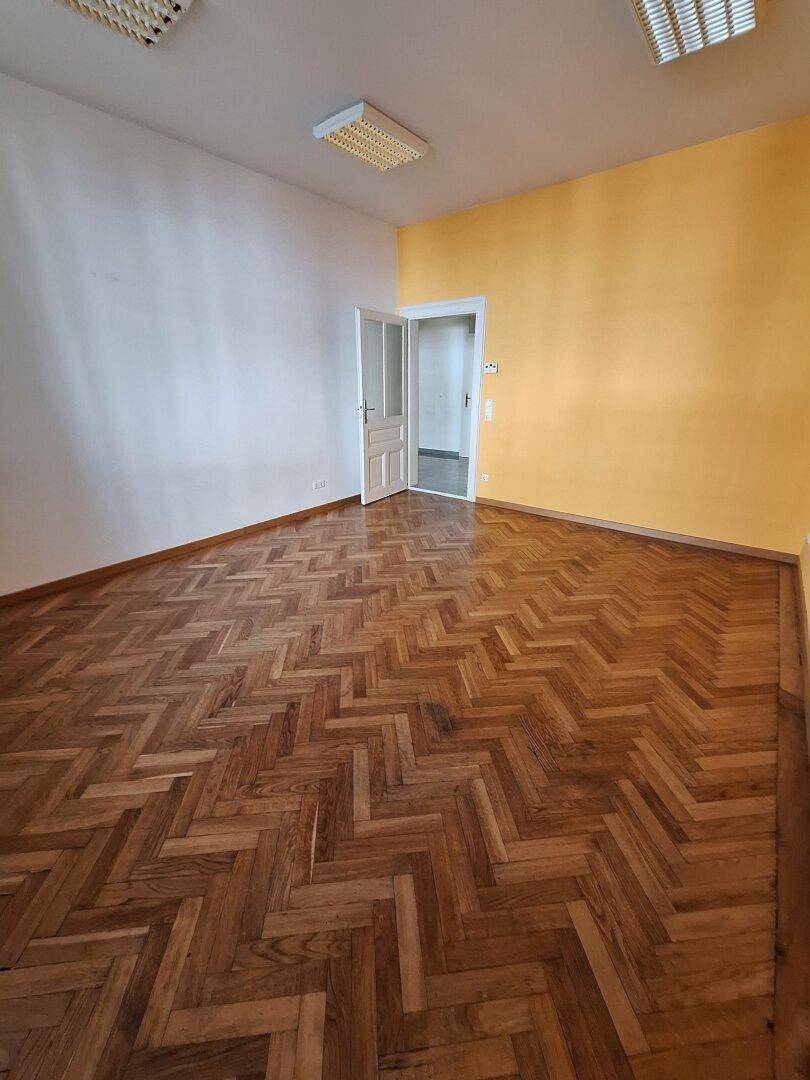 Heller Raum mit Fischgrätparkett, einer gelben Akzentwand und einer offenen Tür zu einem weiteren Bereich.