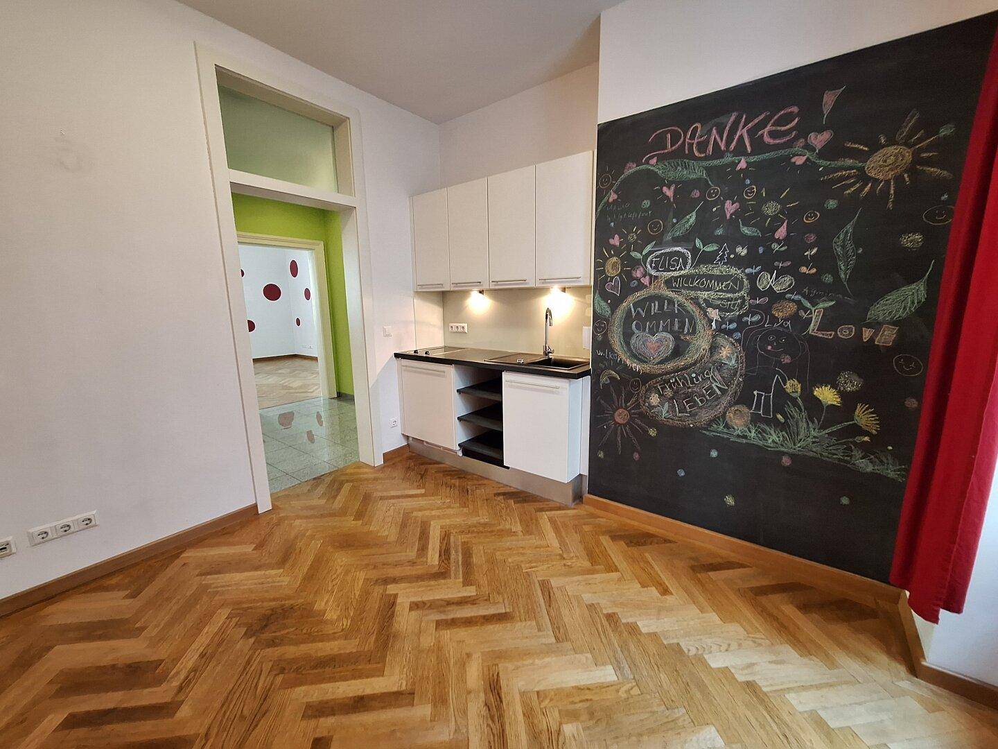 Küchenzeile mit weißen Schränken, schwarzer Arbeitsplatte und Kreidetafelwand, mit Blick in einen grünen Raum.
