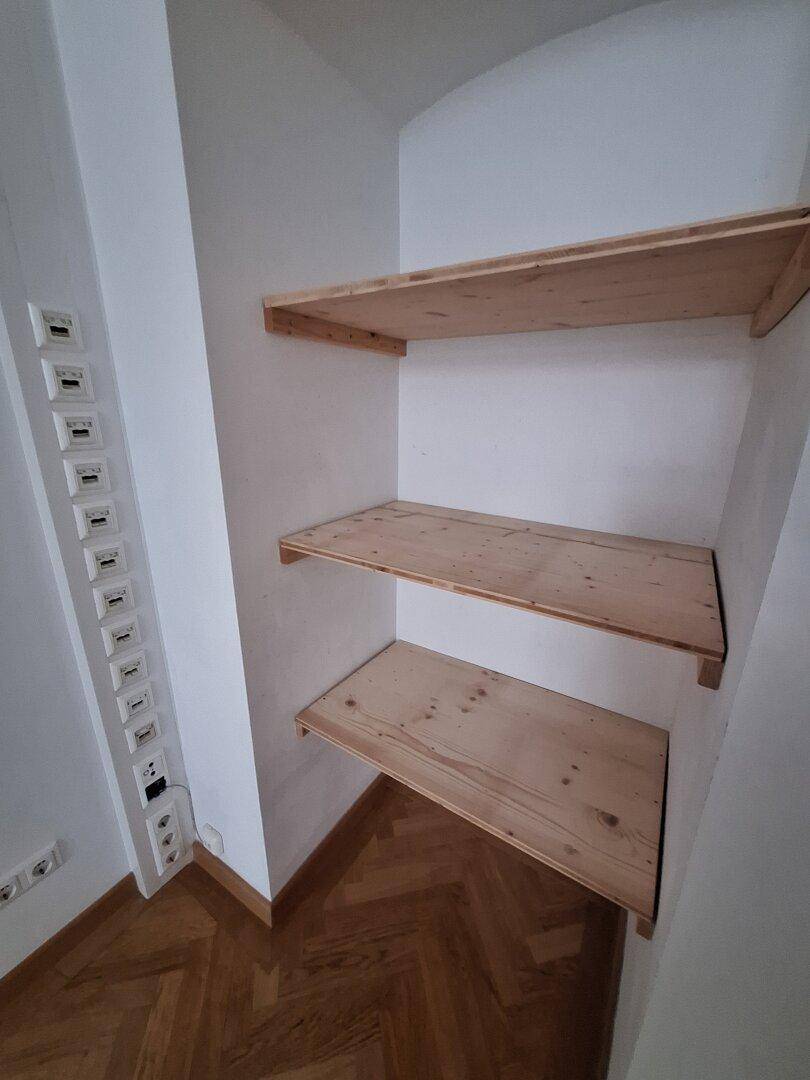 Kleiner Abstellraum mit weißen Wänden und drei einfachen Holzregalen über Fischgrätparkett.
