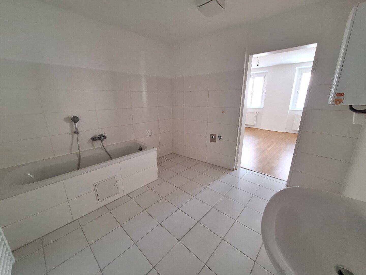 Badezimmer mit weißen Fliesen, Badewanne und Waschbecken, mit Blick in einen angrenzenden Raum.