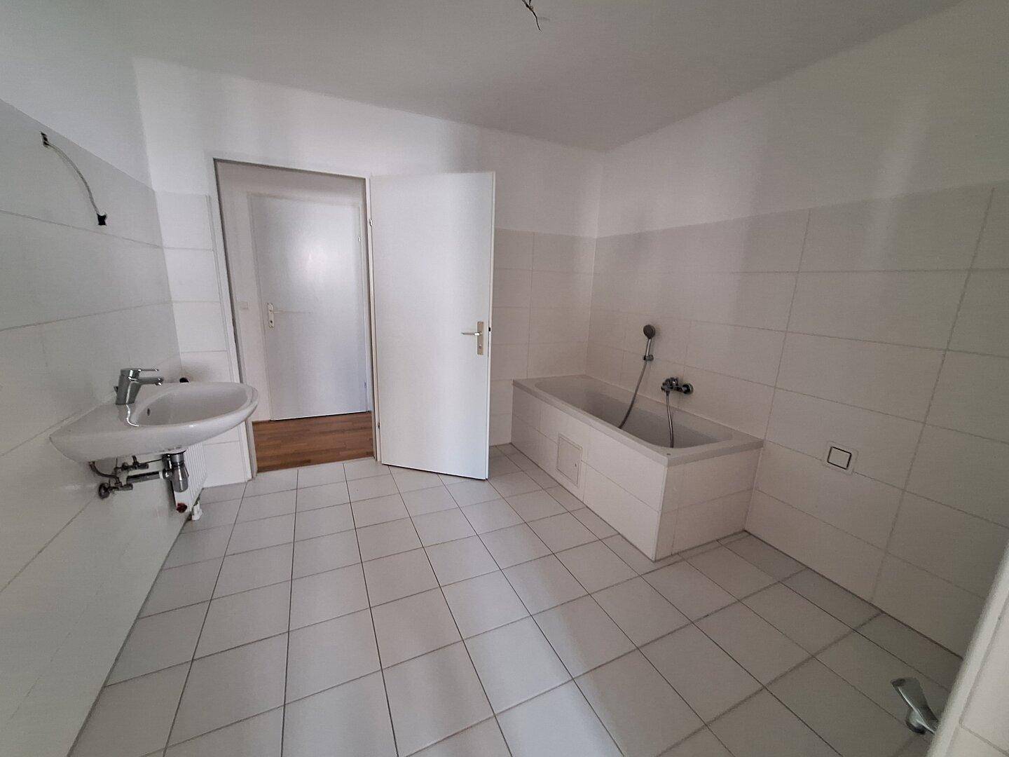 Weiß gefliestes Badezimmer mit Badewanne, Waschbecken und einer Tür, die zu einem anderen Raum führt.