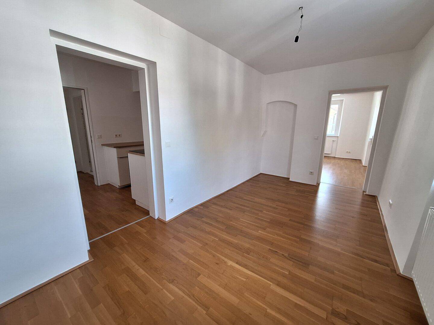 Heller Raum mit Parkettboden, der zu einer offenen Küche und weiteren Zimmern führt.