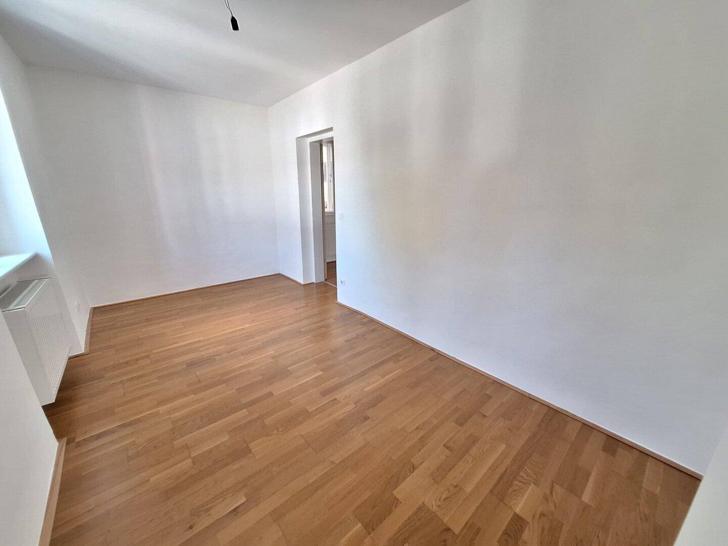Leeres, helles Zimmer mit Parkettboden und einem Fenster, das viel Licht spendet.