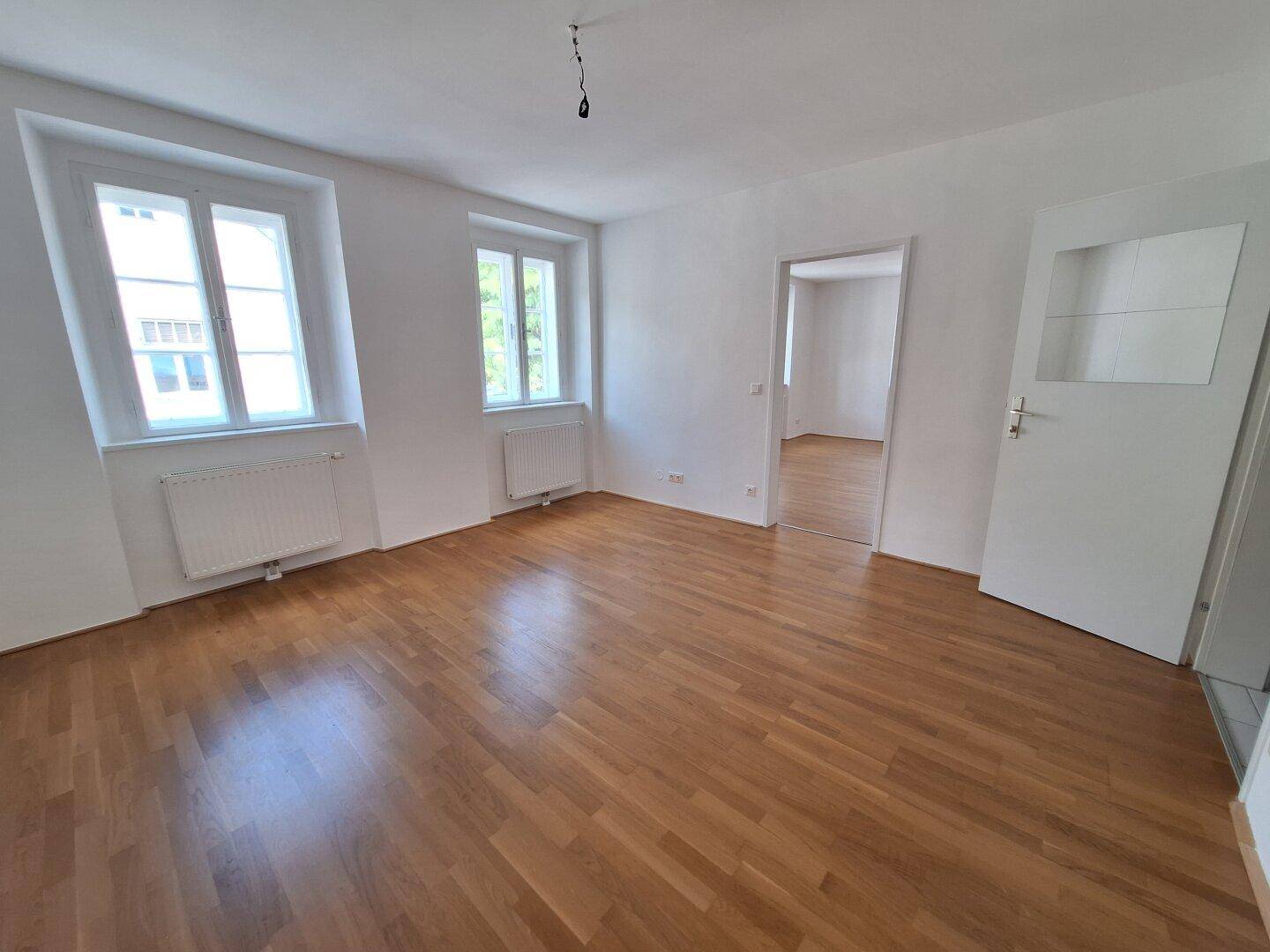 Freundliches Zimmer mit Holzboden, zwei Fenstern und einer Tür mit Glaseinsatz.