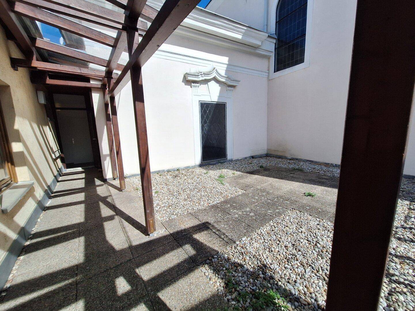 Außenbereich mit Kies und Steinplatten unter einer Holzpergola, die Schatten spendet.