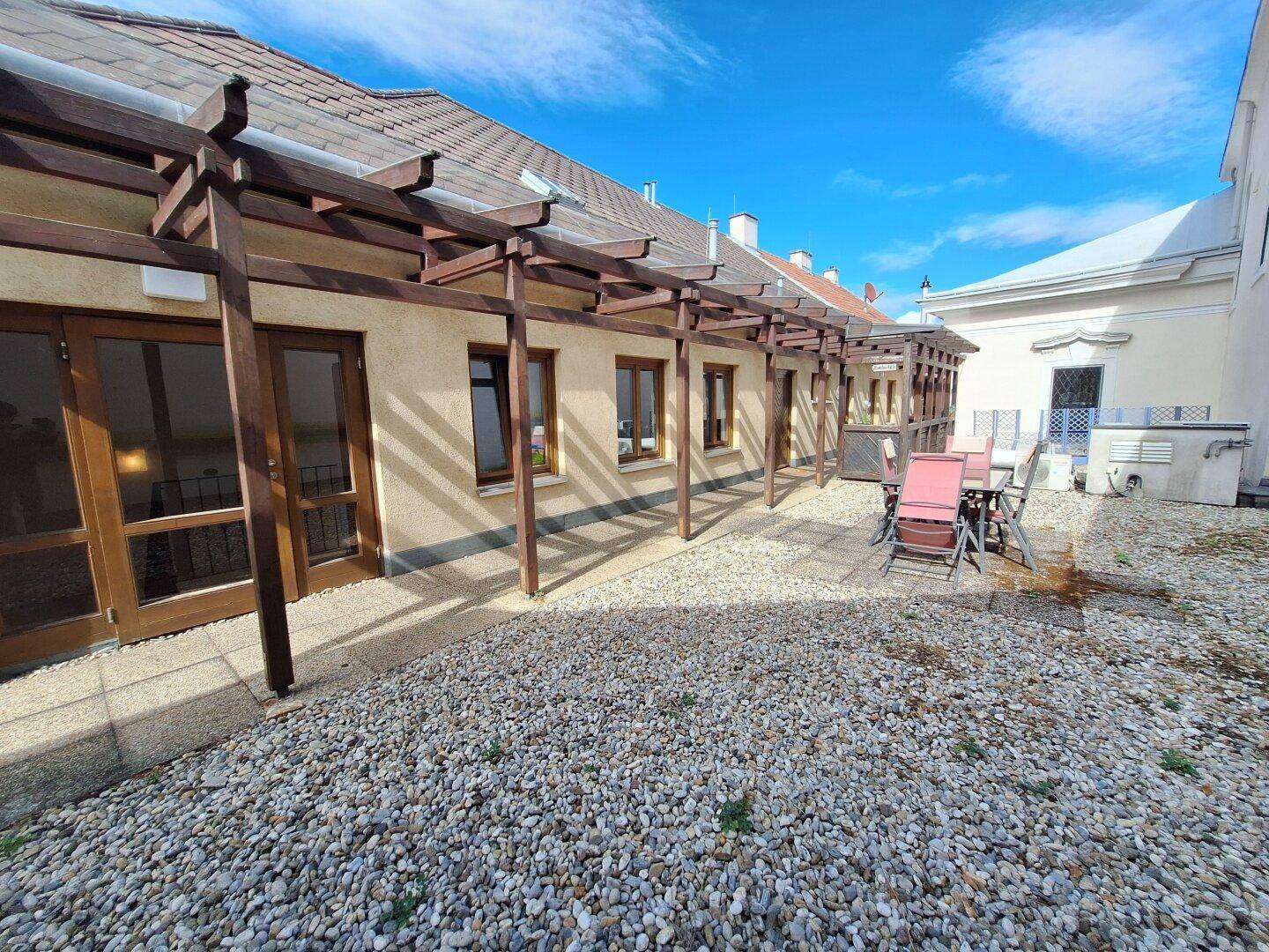 Große Terrasse mit Kies und Gartenmöbeln, umgeben von einer Holzpergola und Gebäuden.