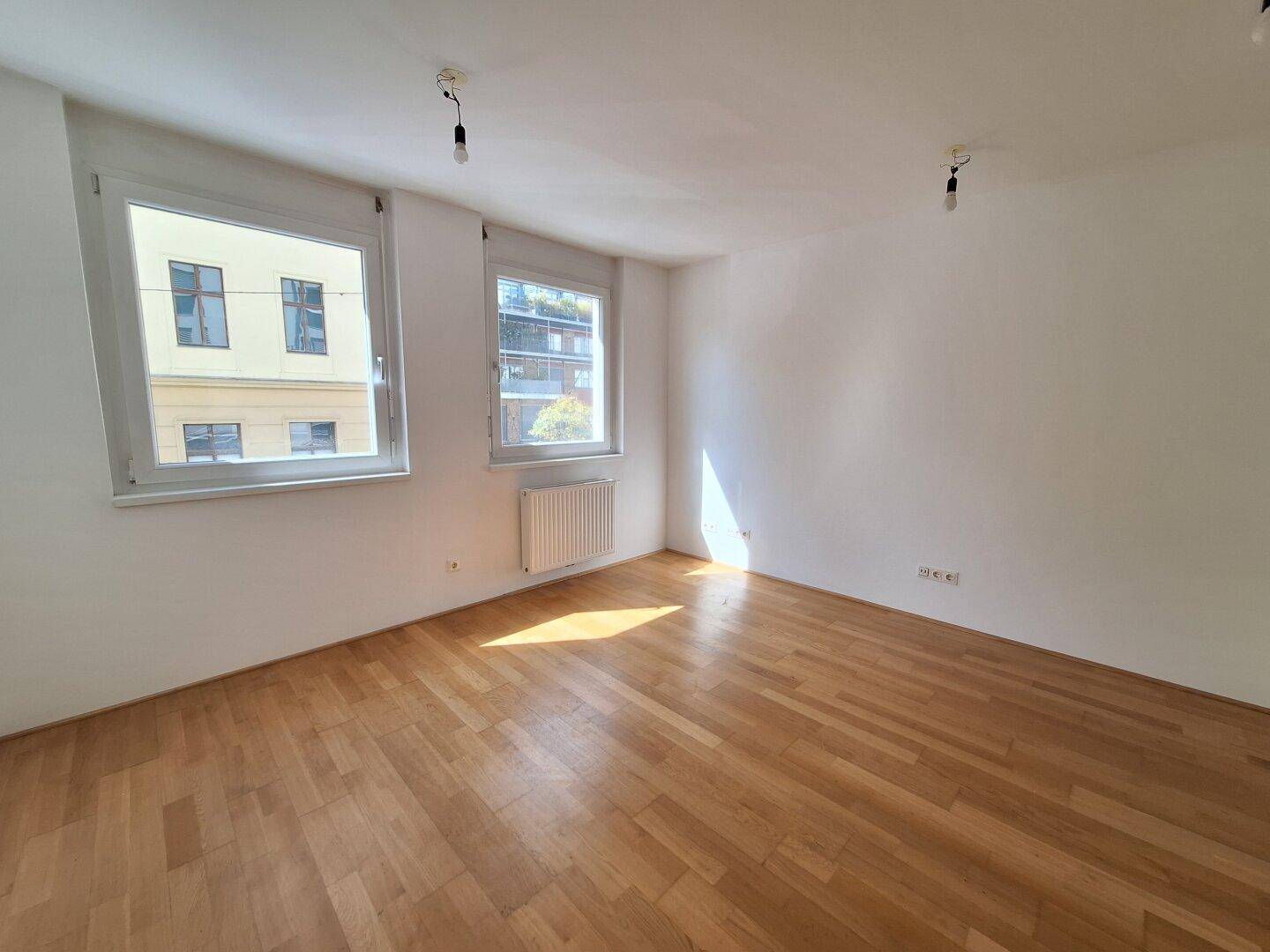 Leeres Zimmer mit Parkettboden, großen Fenstern und Heizkörper unter dem Fenster.