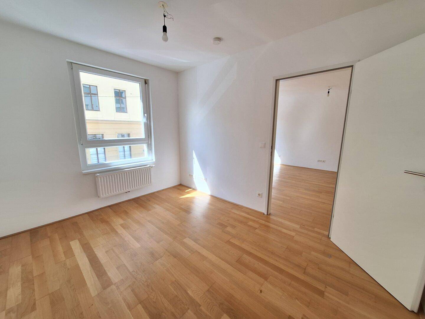 Helles Schlafzimmer mit Parkettboden, Fenster und Zugang zu einem weiteren Raum.