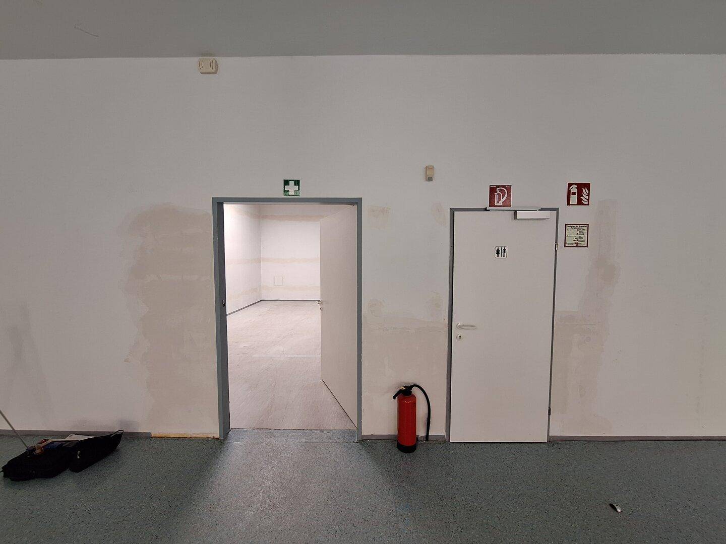 Flur mit zwei Türen, einem Feuerlöscher und Hinweisschildern in einem gewerblichen Gebäude.