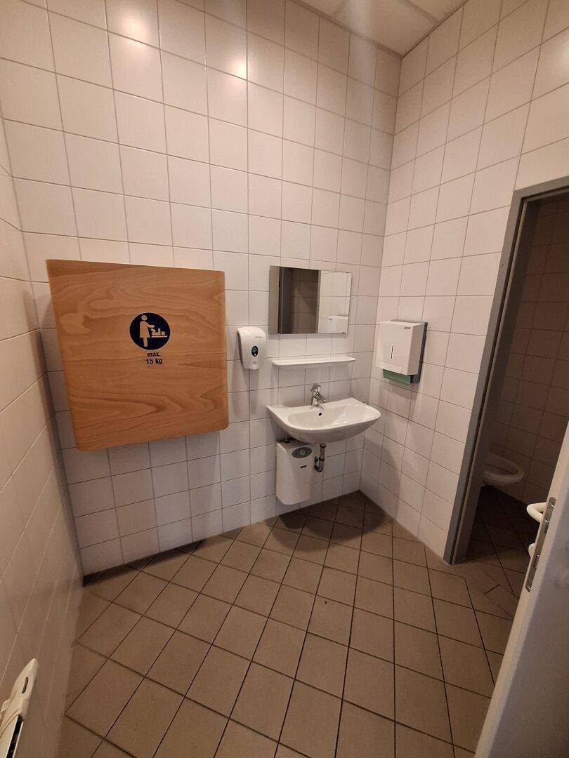 Gefliester Toilettenraum mit Waschbecken, Spiegel und Wickeltisch an der Wand.
