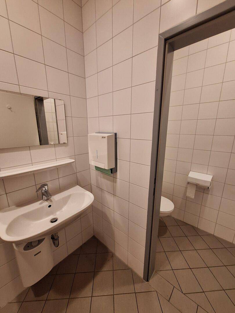 Gefliester Toilettenraum mit Waschbecken, Spiegel und Papierhandtuchspender.