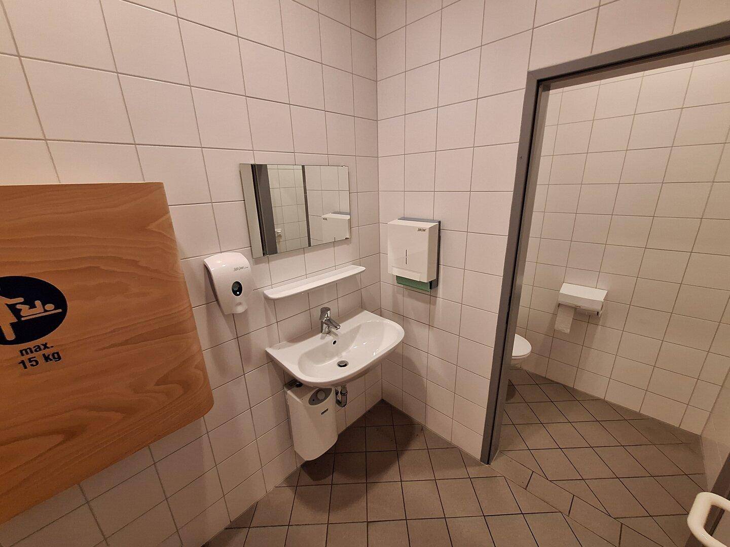Badezimmer mit weißen Fliesen, Waschbecken, Spiegel und Wickeltisch-Symbol, Toilette im Hintergrund sichtbar.
