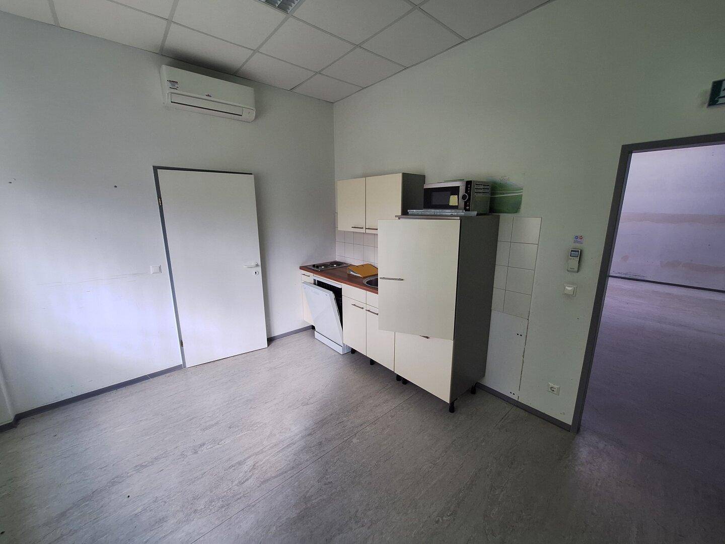 Kleine Küchenzeile mit Spüle, Kochfeld, Kühlschrank und Mikrowelle in einem Büroraum.