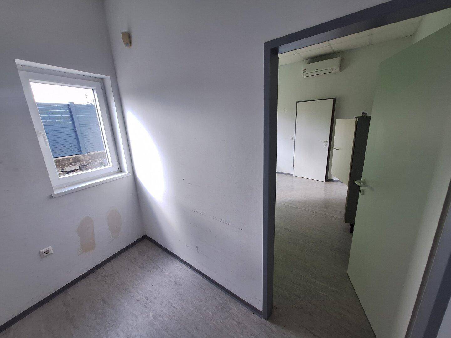 Kleines Büro mit weiß gestrichenen Wänden, einem Fenster und einer Tür, die zu einem weiteren Raum führt.