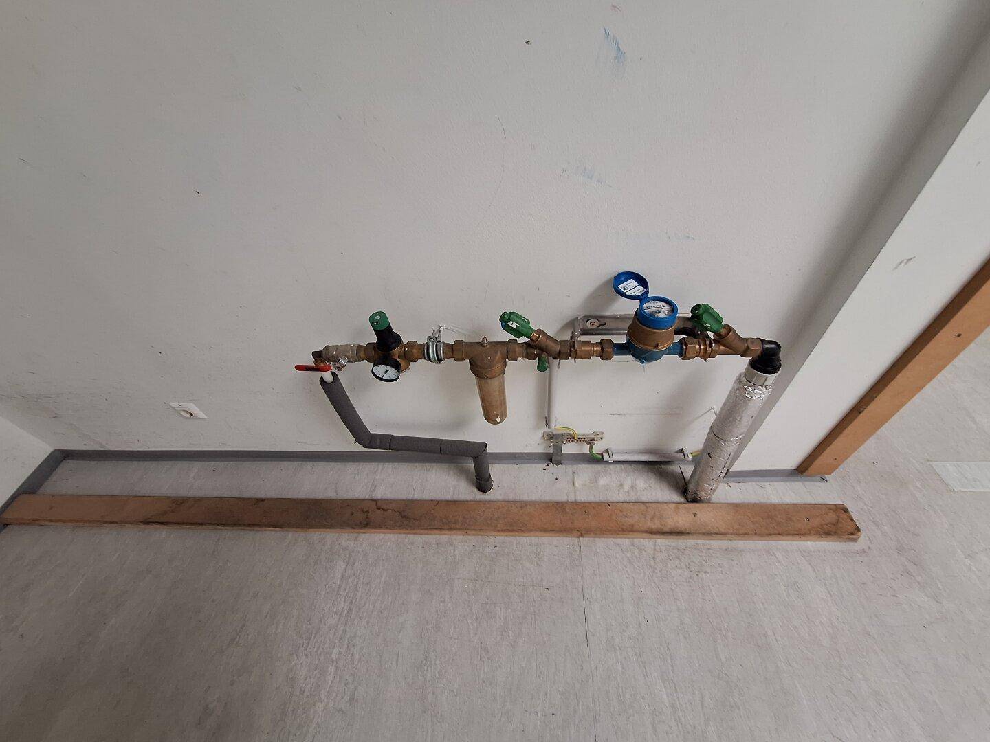 Freiliegende Wasserleitungen und Messgeräte an einer weißen Wand in einem Technikraum.