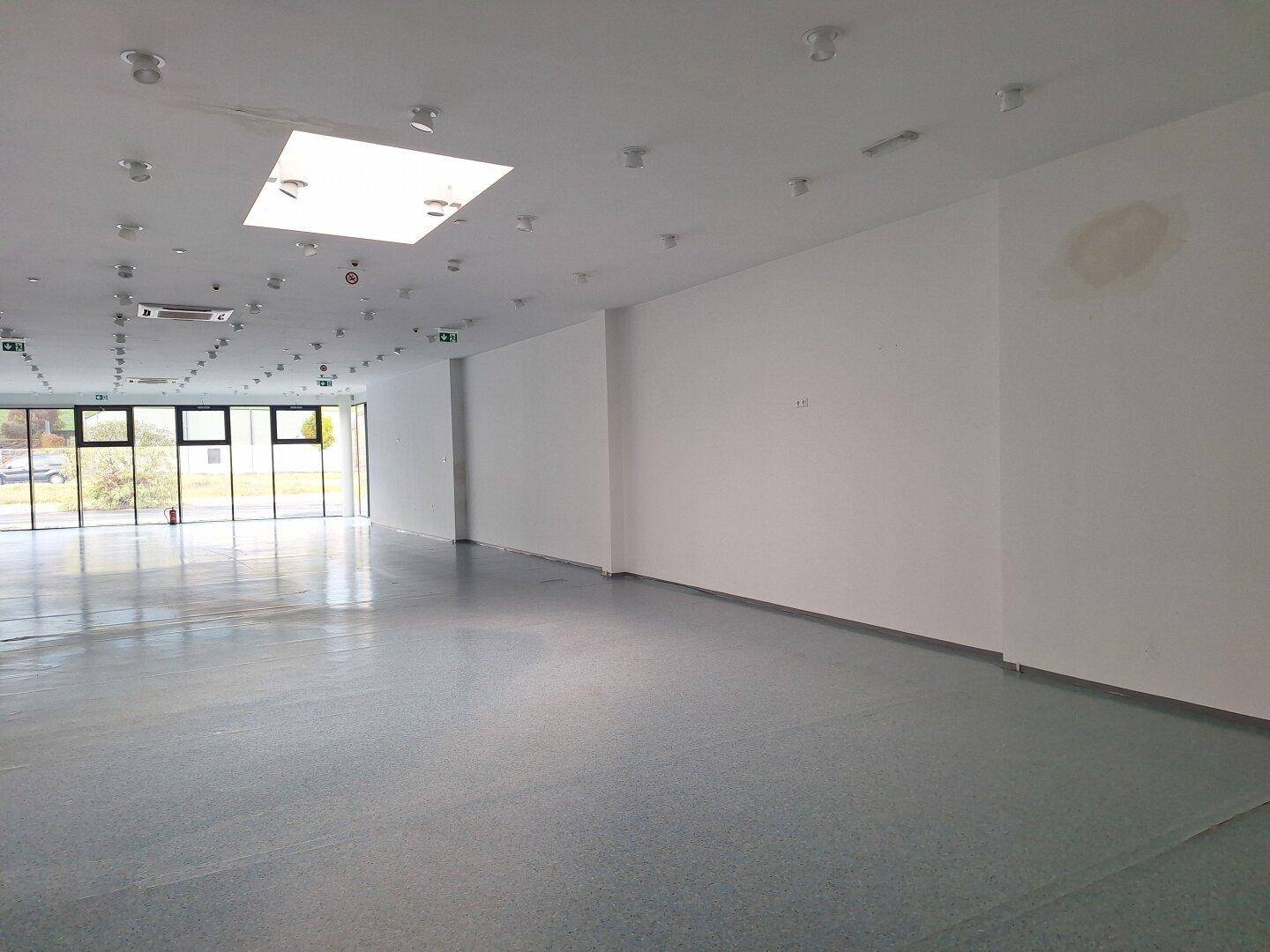 Heller Gewerberaum mit großen Fensterfronten und einer weißen Wand, die leichte Gebrauchsspuren aufweist.