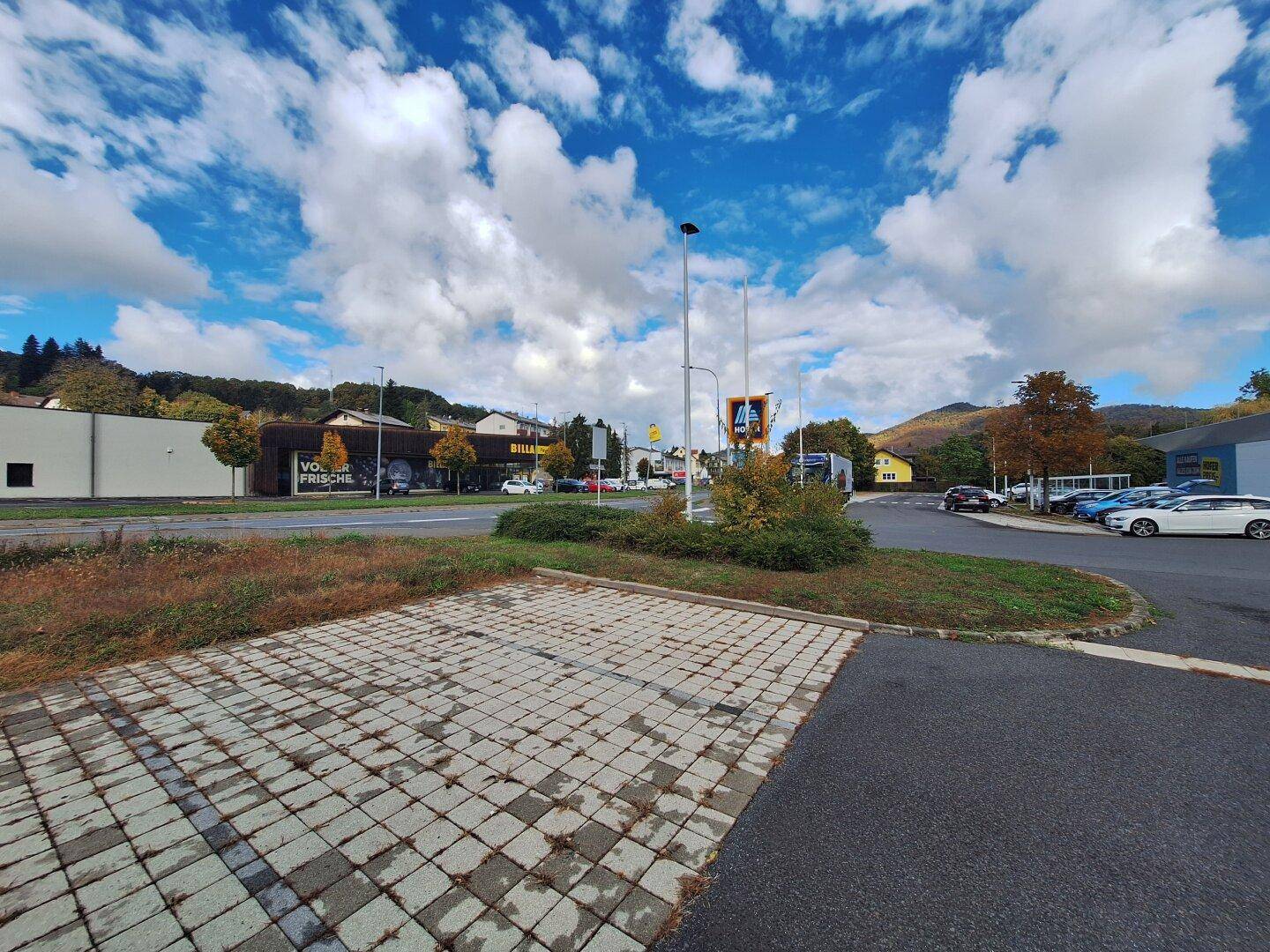 Außenansicht eines Parkplatzes mit gepflasterten Flächen und einem Supermarkt im Hintergrund unter blauem Himmel.