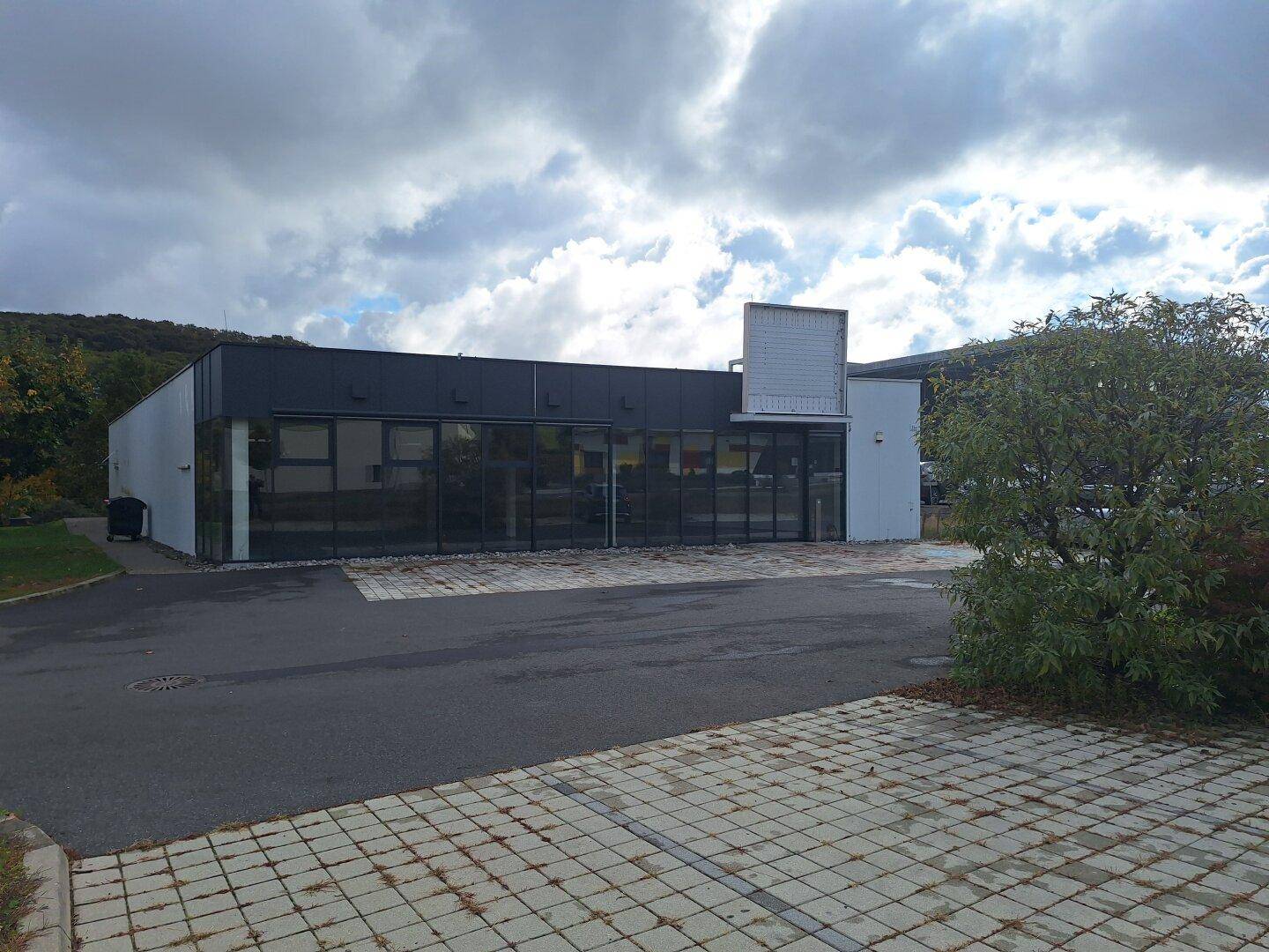 Moderne Geschäftsfassade mit großen Glasfenstern und einem Parkplatz davor unter bewölktem Himmel.