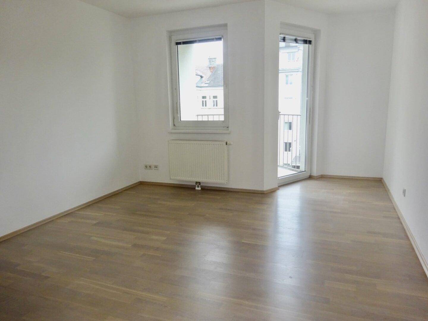 Leeres Zimmer mit Parkettboden und Zugang zu einem französischen Balkon.