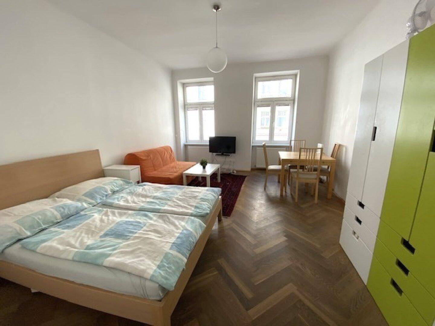 Helles Zimmer mit Schlafbereich, Esstisch und Sofa, ausgestattet mit Parkettboden und großen Fenstern.