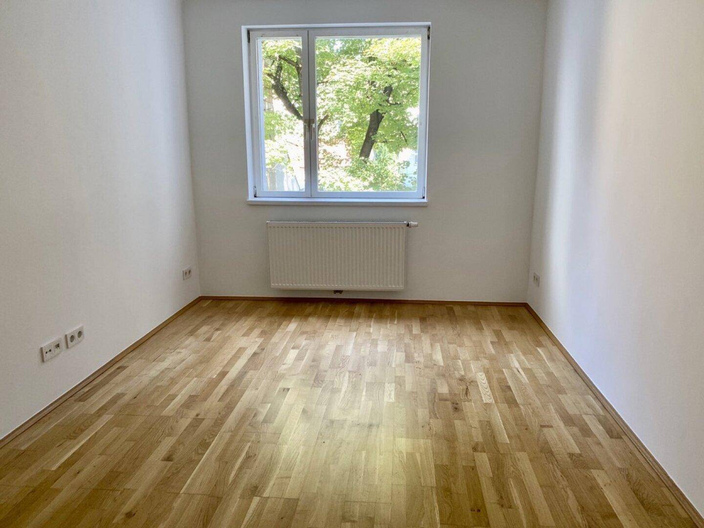 Heller Raum mit Parkettboden und Fenster mit Blick ins Grüne.
