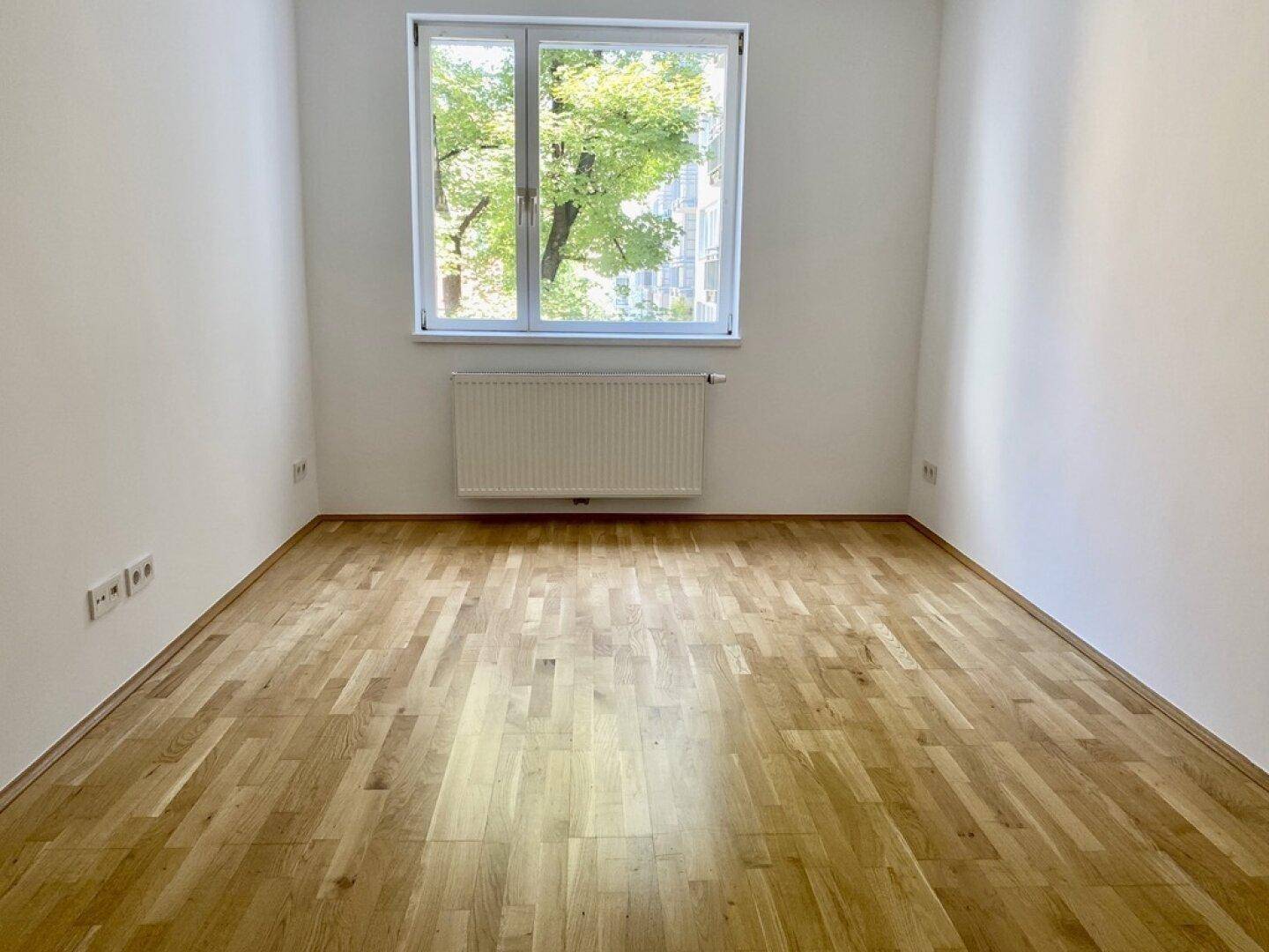 Geräumiger Raum mit Parkettboden und großem Fenster mit Blick ins Grüne.
