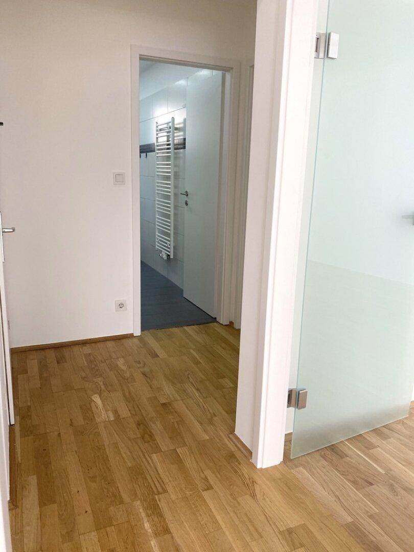 Heller Flur mit Parkettboden und Zugang zum Badezimmer durch eine Glastür.