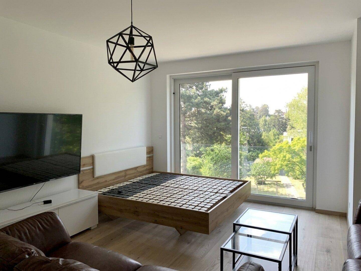 Helles Schlafzimmer mit Holzbettrahmen und großem Fenster mit Blick ins Grüne.