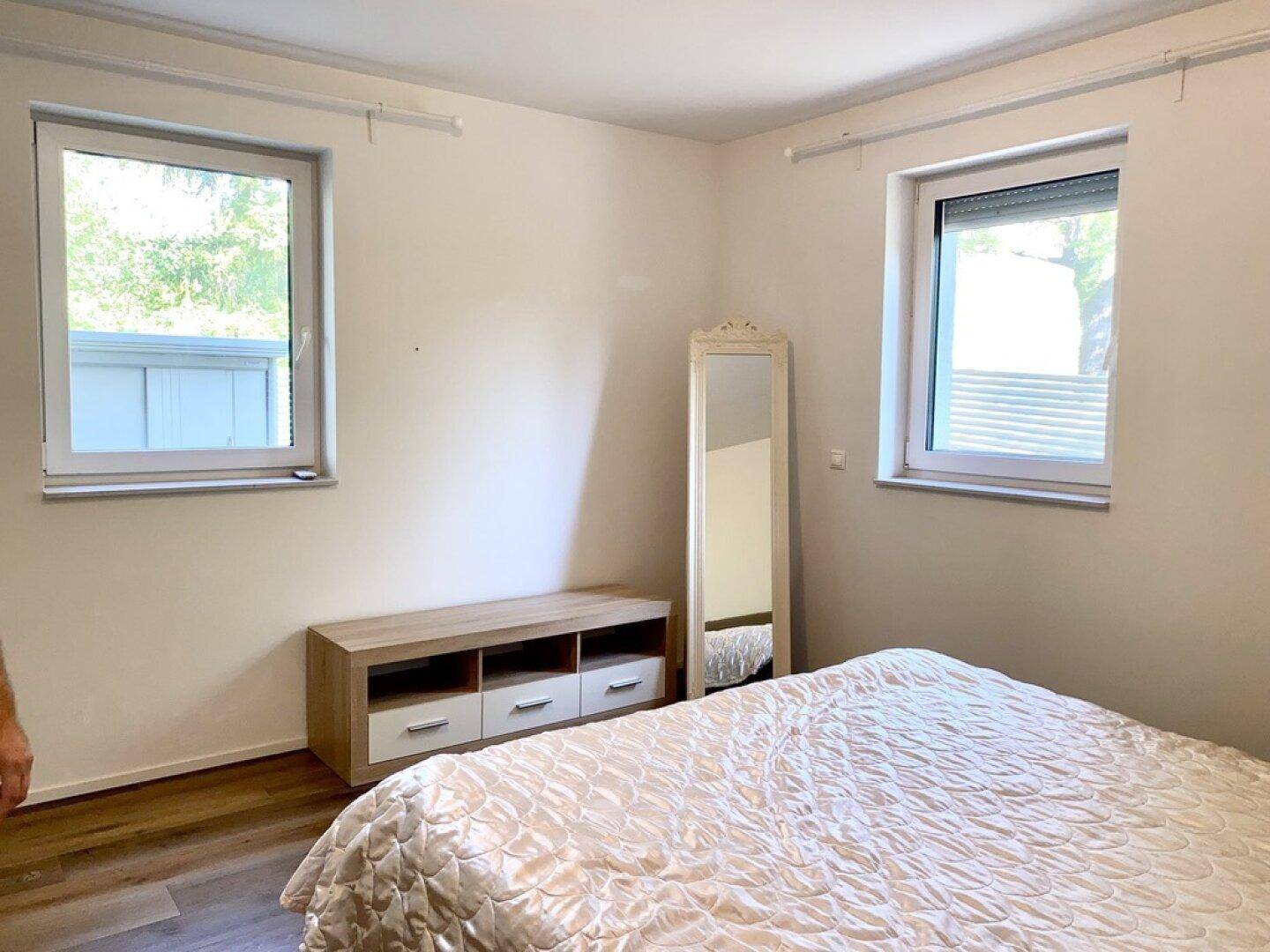 Helles Schlafzimmer mit Doppelbett, Kommode und zwei Fenstern mit Rollläden.