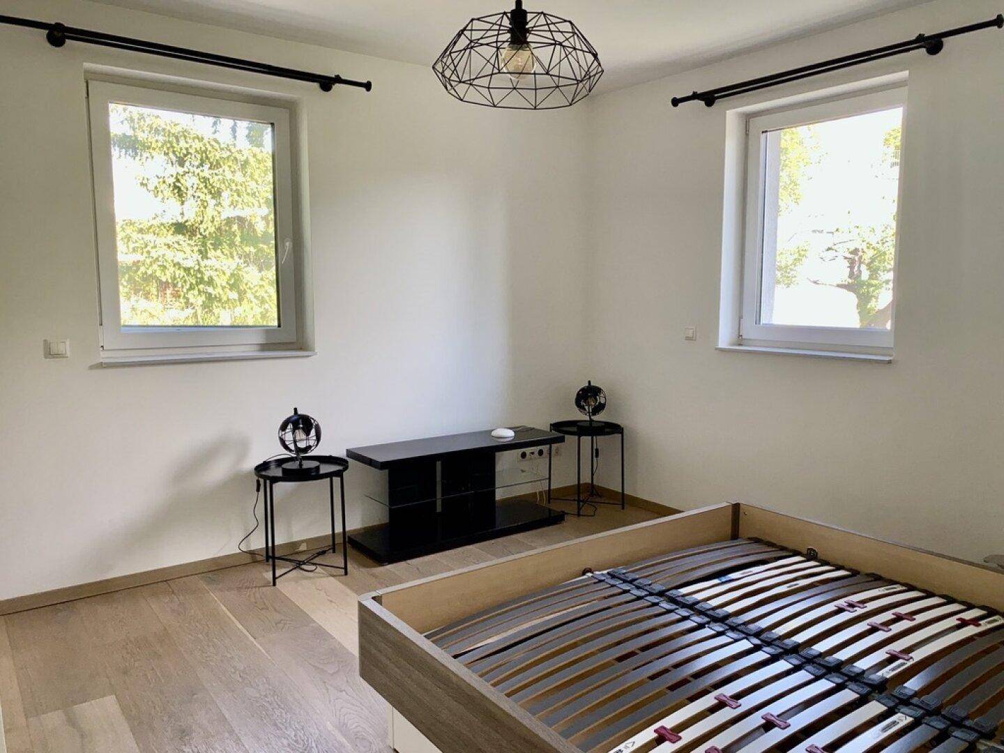 Helles Schlafzimmer mit modernem Bettrahmen und zwei Fenstern, die viel Licht spenden.