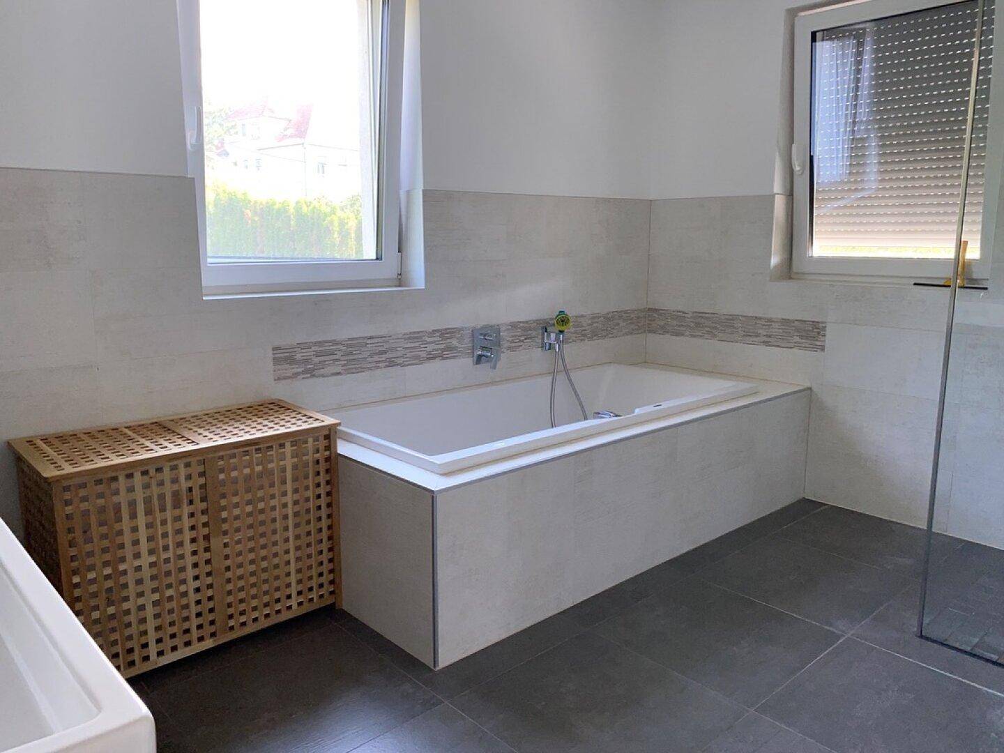 Helles Badezimmer mit eingebauter Badewanne, zwei Fenstern und modernen Fliesen.