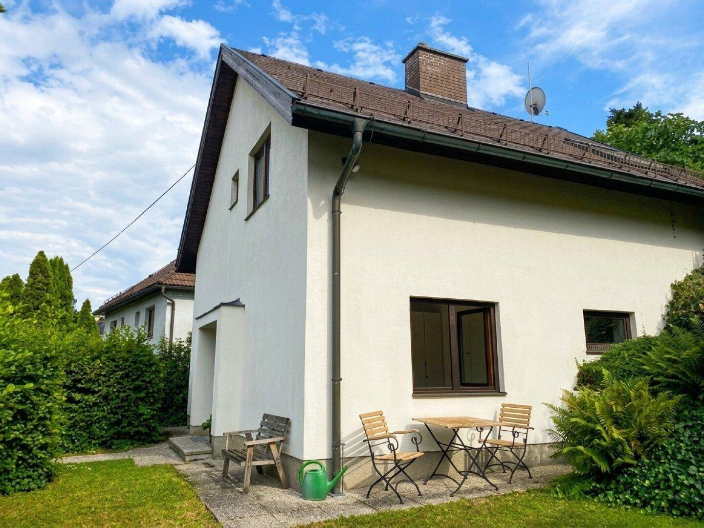 Seitenansicht des Hauses mit weißer Fassade, braunen Fenstern und einem kleinen Sitzbereich im Freien.