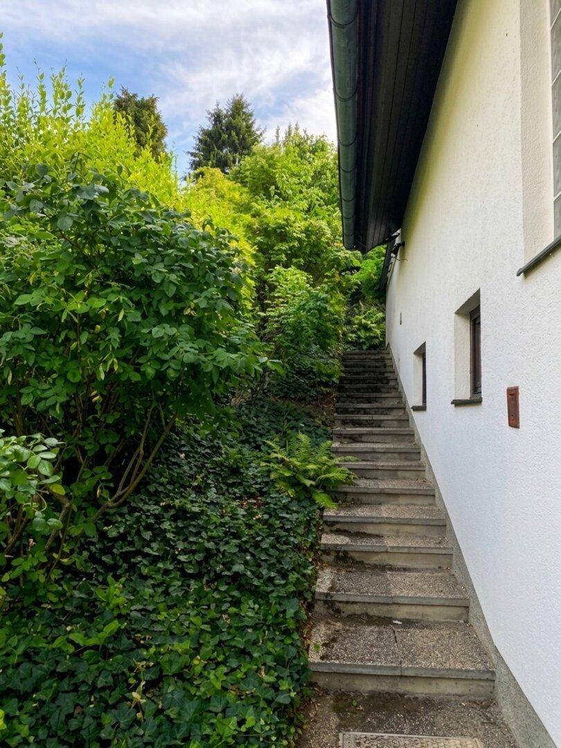 Steile Steintreppe führt einen bewachsenen Hang hinauf, neben der weißen Hauswand.