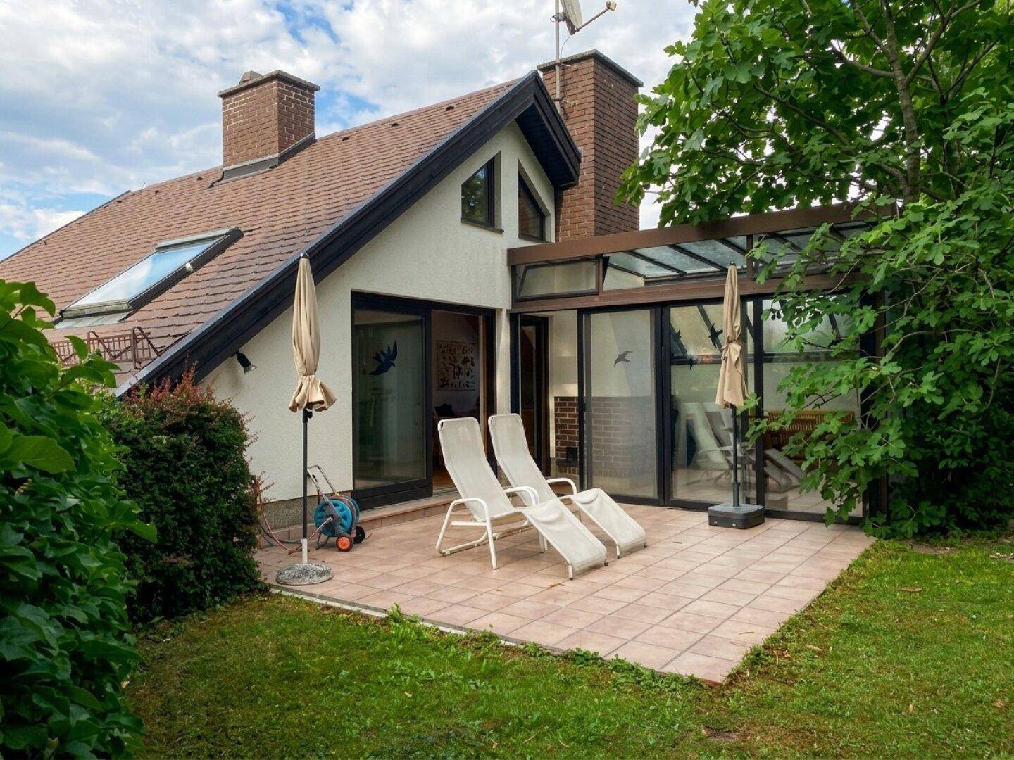 Große gepflasterte Terrasse mit Liegestühlen und Sonnenschirmen vor einem Wintergarten und dem Haus.