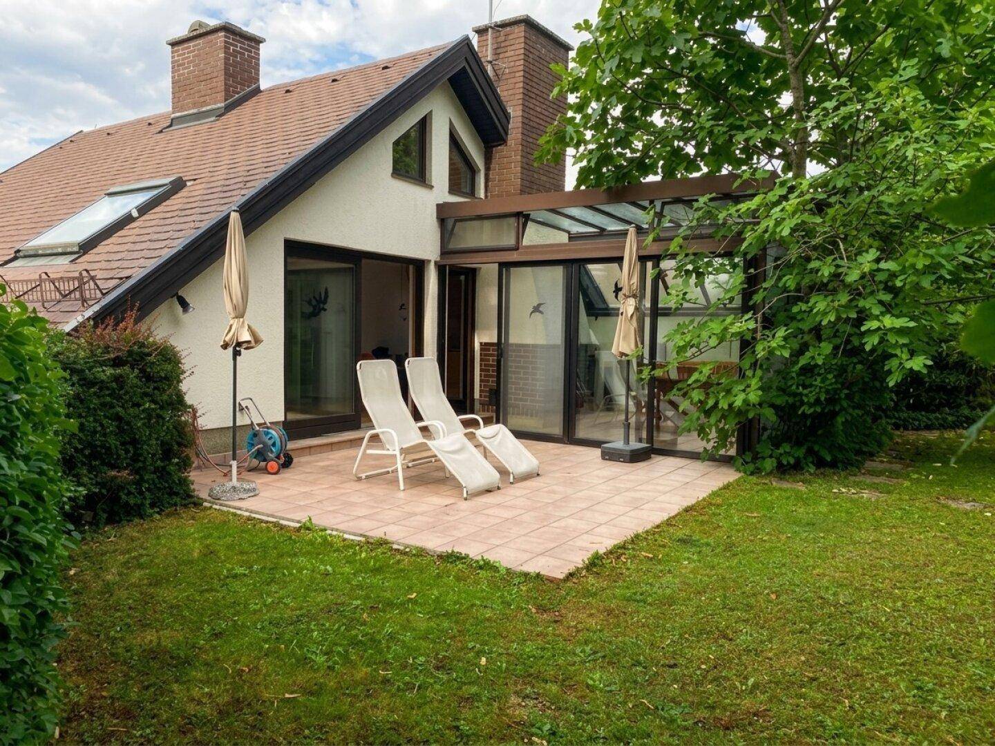 Rückseite des Hauses mit einer gepflasterten Terrasse, Sonnenliegen und einem Wintergarten.