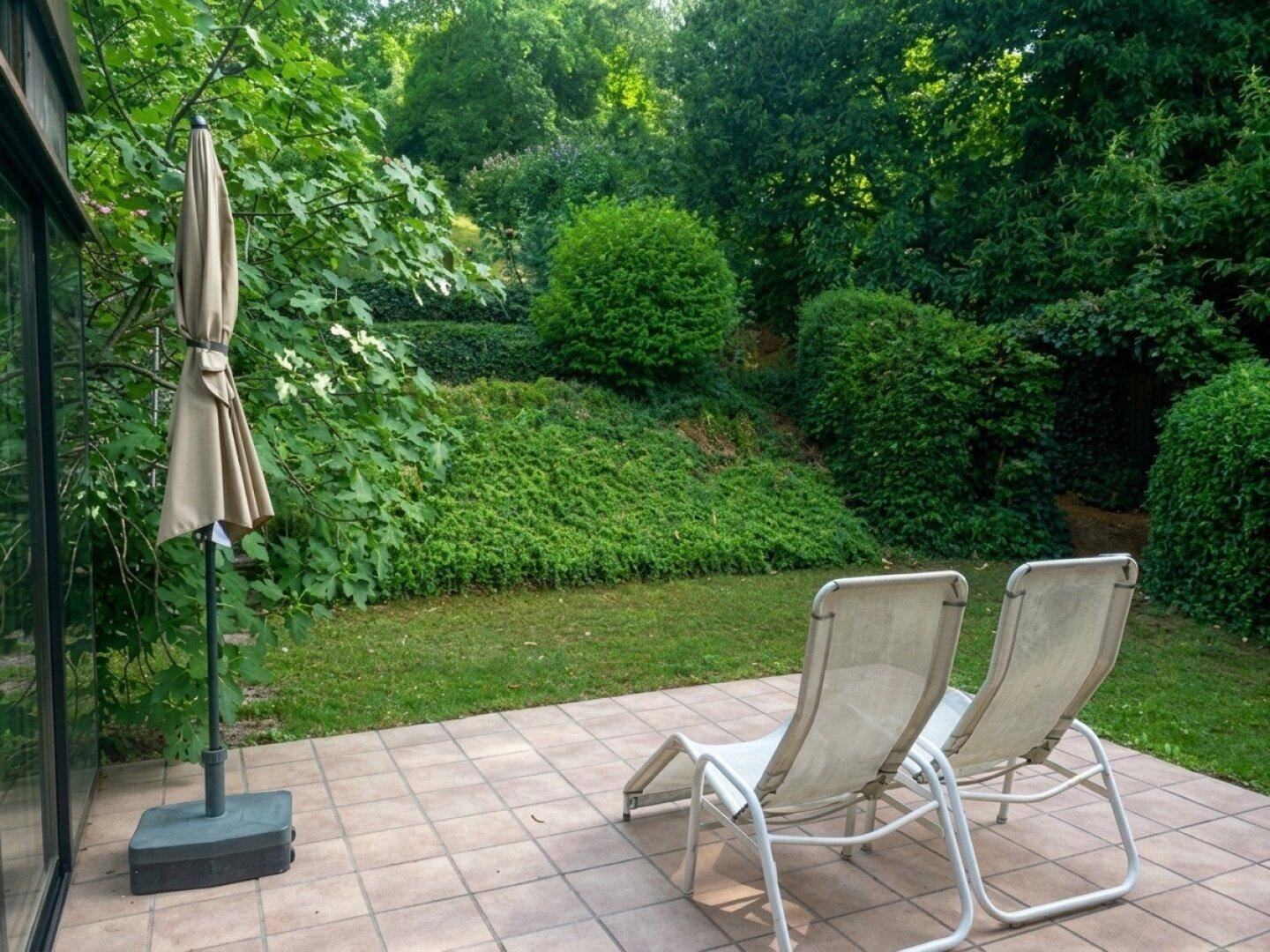 Gepflasterte Terrasse mit Liegestühlen und Sonnenschirm, umgeben von dichtem Grün und Bäumen.