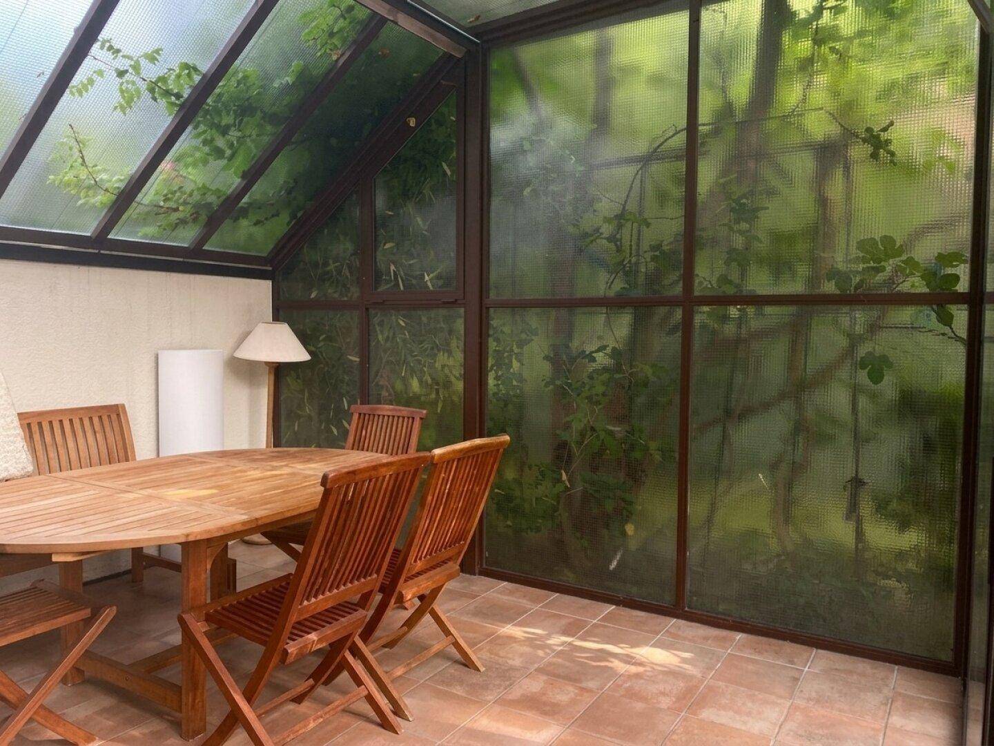 Heller Wintergarten mit Holztisch und Stühlen, umgeben von üppigem Grün durch die Glaswände.