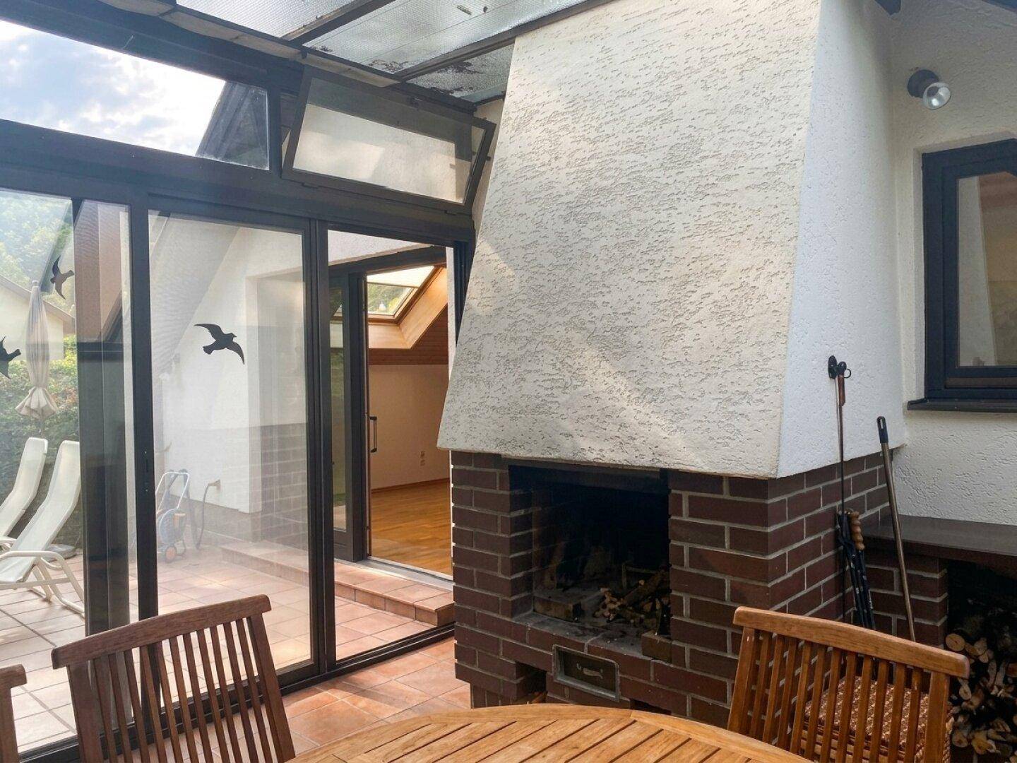 Gemütlicher Wintergarten mit gemauertem Kamin, Holztisch und Blick auf die Terrasse.